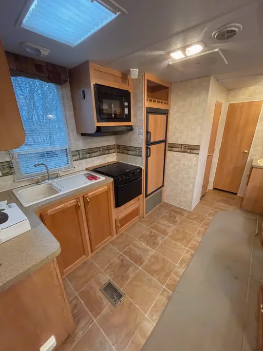 This bright RV rental kitchen features a window, sink, stove, microwave, fridge, and wood-look cabinets, ideal for road trips.. Forest River Cherokee 2008