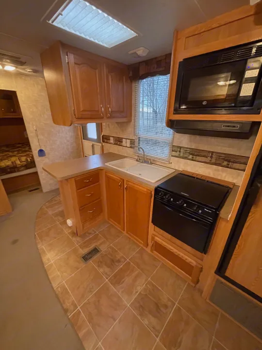 This RV rental kitchen features wood cabinets, a double sink, black microwave, stove, and tiled flooring.. Forest River Cherokee 2008