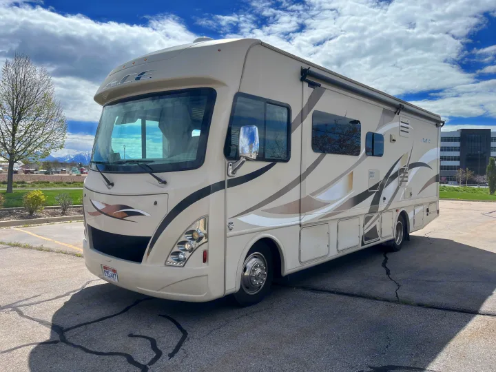 This large beige RV motorhome, available for rv rental, is parked on asphalt with mountains and trees in the background.. Thor A.C.E. 2016