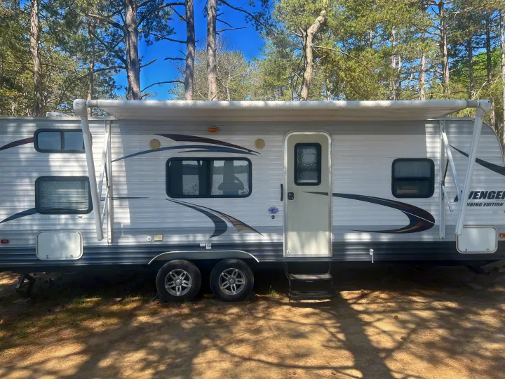 A spacious white and gray rv rental travel trailer with dual axles and an extended awning is in a sunny, wooded campsite.. Forest River Wildwood X-Lite 2023