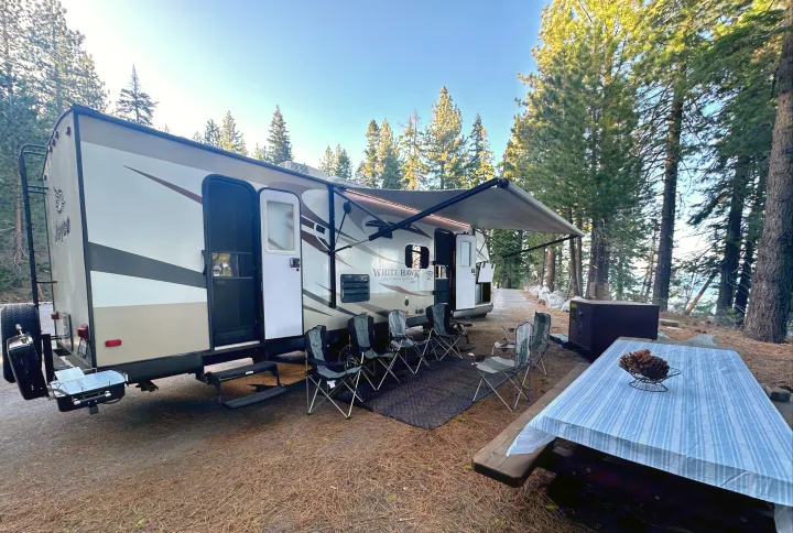 An RV rental travel trailer at a serene campsite with an open awning, outdoor seating, and a picnic table among tall pines.. Jayco White Hawk Ultra Lite 2015