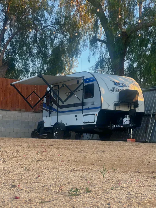 An inviting RV rental travel trailer, with its awning extended, parked on a gravel lot under trees with string lights.. Jayco Hummingbird 2019