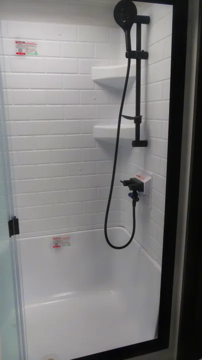 Modern RV rental shower with white subway tile pattern, black fixtures, corner shelves, and clear glass door.. Forest River Rockwood 2025