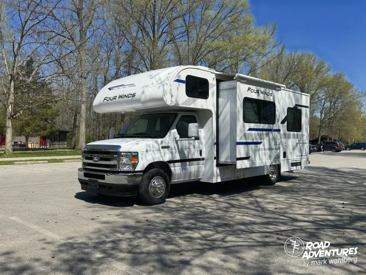 White Four Winds Class C RV rental with slide-out, parked in a sunny lot surrounded by trees. Ready for your adventure.. Thor Motor Coach Four Winds 2026