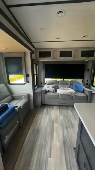 Modern RV rental interior featuring gray couches, wood-look flooring, overhead cabinets, and black-out window blinds.. Keystone Sprinter 2022