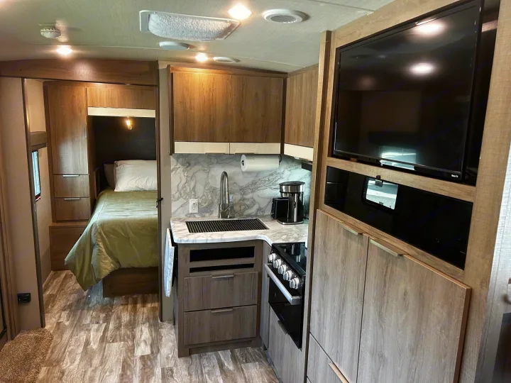 Modern RV rental interior: compact kitchen with stove, bed, TV, & wood-look cabinetry.. Grand Design Imagine 2022