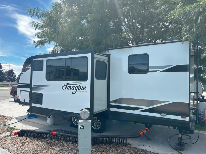 A white Imagine travel trailer RV rental is parked at a campsite with its slide-out extended, connected to hookups for a stay.. Grand Design Imagine 2022