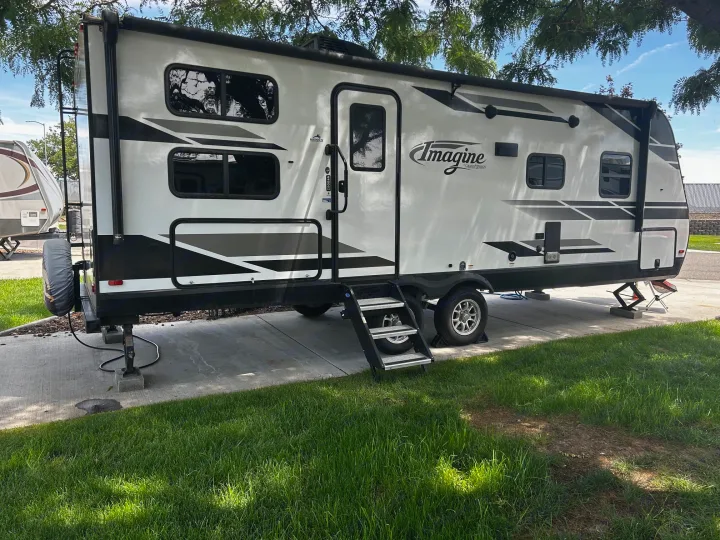 White Imagine travel trailer RV rental, equipped with steps and stabilizers, parked on a concrete pad under a tree.. Grand Design Imagine 2022