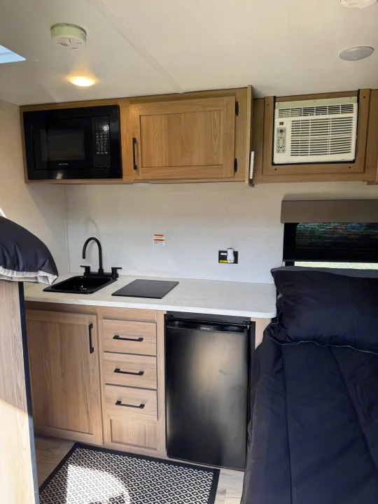 Inside a modern RV rental: compact kitchen with microwave, sink, fridge, and cozy bed area.. Keystone RV Coleman 13B 2025