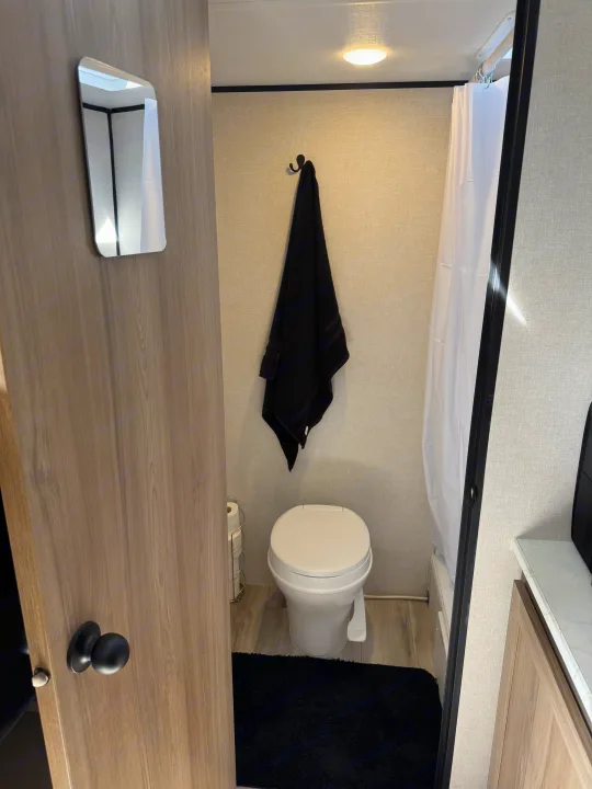 Inside an RV rental bathroom: a white toilet, black towel, shower curtain, and a mirror on the door. Compact and functional.. Keystone RV Coleman 13B 2025