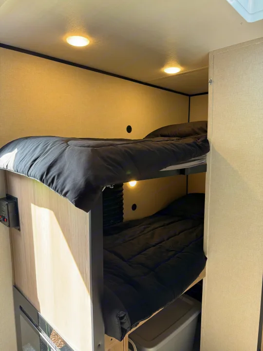 RV rental interior: two bunk beds with black comforters, warm overhead lighting, and light wood-paneled walls.. Keystone RV Coleman 13B 2025