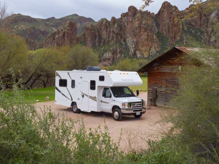 A white Winnebago Minnie Winnie RV rental is parked on a dirt lot beside a rustic building, with rugged mountains behind.. Winnebago Minnie Winnie 2020