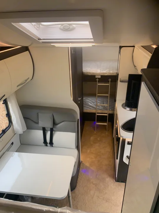 Inside a modern RV rental: a compact interior features a bright dining area, functional kitchen, and cozy bunk beds at the rear.. Rollerteam 2021