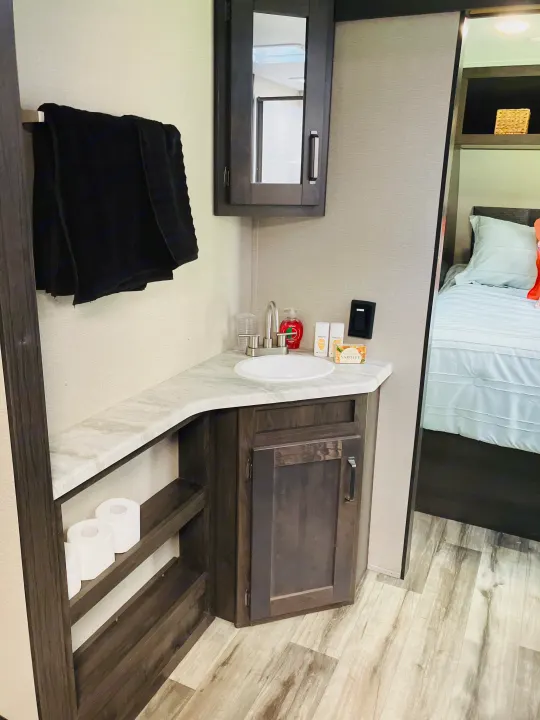 Compact RV rental interior featuring a functional bathroom vanity with sink and storage, adjacent to a cozy bedroom area.. Grand Design 265BH 2023