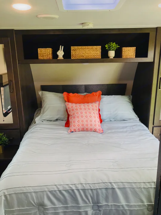 Cozy RV rental bedroom featuring a comfortable bed with light blue bedding, orange decorative pillows, and overhead storage shelves.. Grand Design 265BH 2023