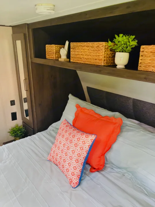 Cozy RV rental bedroom featuring a bed with coral and patterned pillows, overhead storage, and decorative plant. Perfect for travel!. Grand Design 265BH 2023