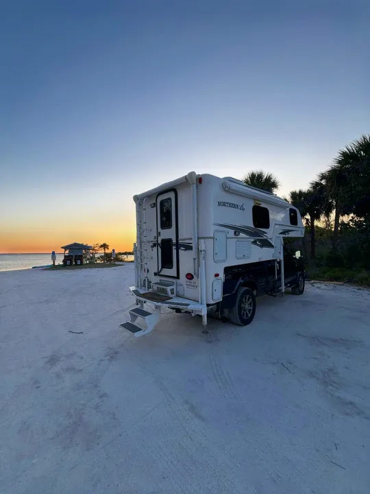 White truck camper RV rental parked on a sandy beach, enjoying a beautiful sunset over the water.. Northern Lite 9-6 Wet Bath 2022
