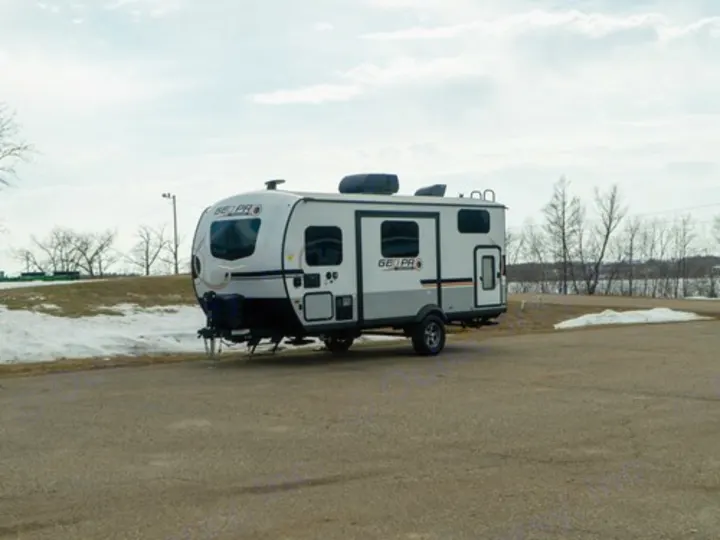 White & gray travel trailer for an RV rental adventure, parked on asphalt. Some snow on ground, trees, and water in the background.. Rockwood GeoPro 2022