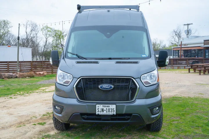 Gray Ford Transit rv rental van with roof rack, front view, parked on gravel/grass by outdoor lights & buildings.. Ford Transit Custom 2022