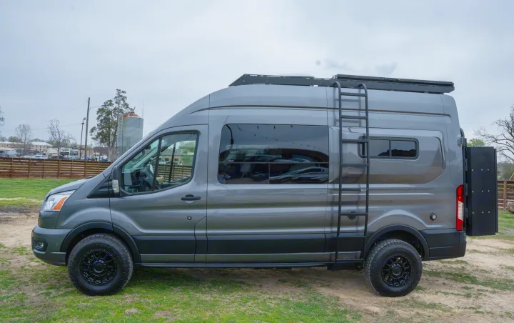 This sleek grey RV rental camper van, equipped with off-road tires, roof rack, and ladder, awaits your adventure.. Ford Transit Custom 2022