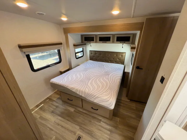 Cozy RV rental bedroom featuring a patterned mattress, tufted headboard, and wood-look flooring, with windows and overhead storage.. Forest River Rockwood Ultra Lite 2024