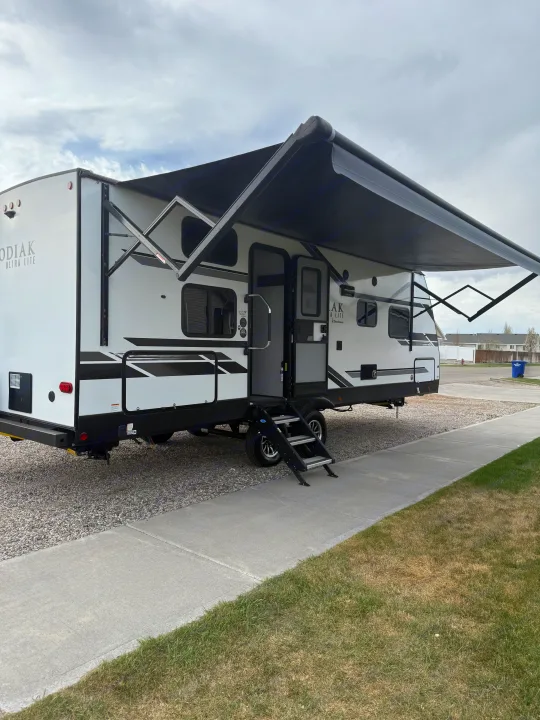 White Kodiak Ultra Lite RV with extended black awning, parked on gravel beside a sidewalk. Perfect for an rv rental.. Dutchmen Kodiak Ultra Lite 2021