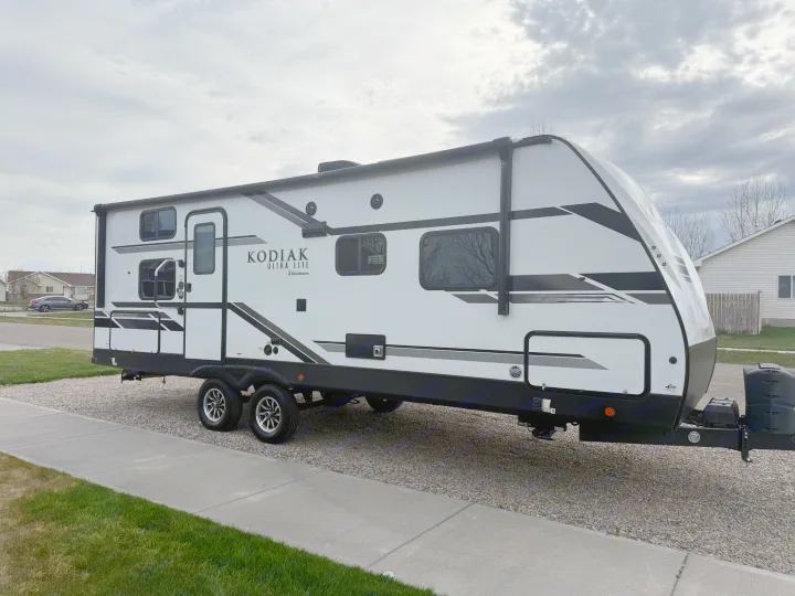 New Kodiak Ultra Lite RV rental, a sleek white travel trailer parked on a gravel lot under an overcast sky.. Dutchmen Kodiak Ultra Lite 2021
