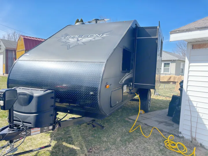 A dark gray Falcon travel trailer RV, connected via a yellow power cord, parked on grass. Perfect for rv rental trips.. Travel Lite Falcon 2018