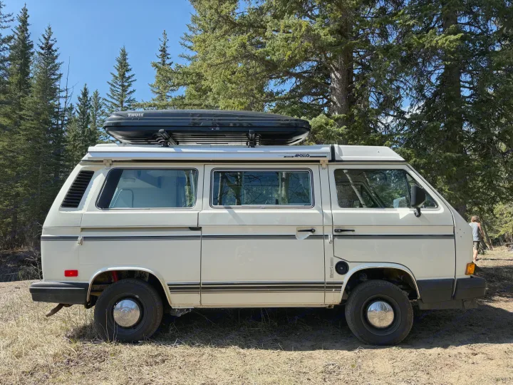 White VW Westfalia camper van with a Thule roof box, parked among pine trees on a clear day. Ideal for an rv rental.. Volkswagen T3 1991