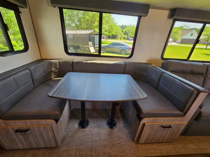 Inside an RV rental: a cozy dinette with grey upholstered benches, wooden table, and outdoor views through large windows.. Keystone RV Passport GT 2022