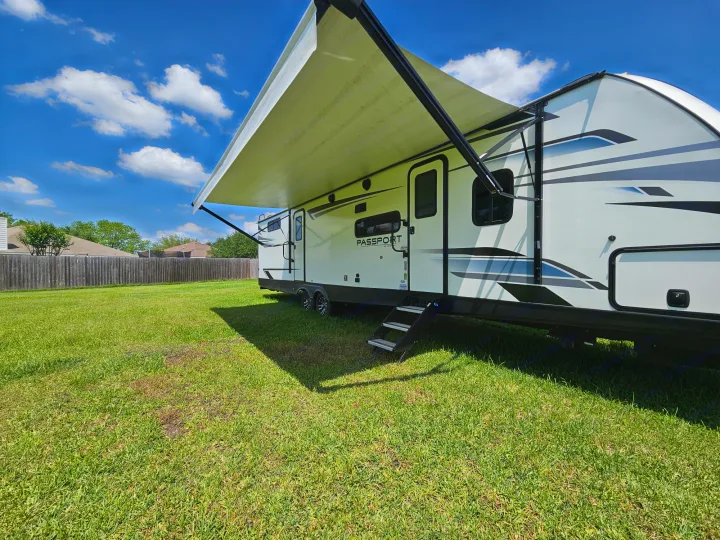 Spacious white RV rental with an extended awning parked on green grass under a sunny blue sky. Ideal for outdoor travel.. Keystone RV Passport GT 2022