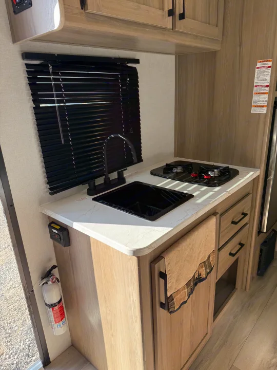 RV rental kitchen interior: functional galley featuring a modern sink, stove, light wood cabinets, and window blinds.. Keystone Coleman 17B 2025