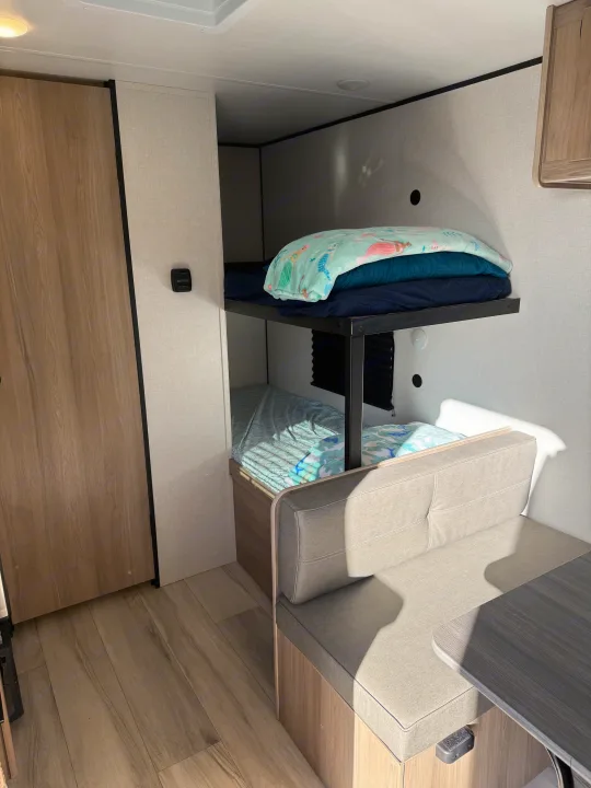 This RV rental features cozy bunk beds with light blue bedding, a tan dinette, and wood-look flooring in a compact space.. Keystone Coleman 17B 2025