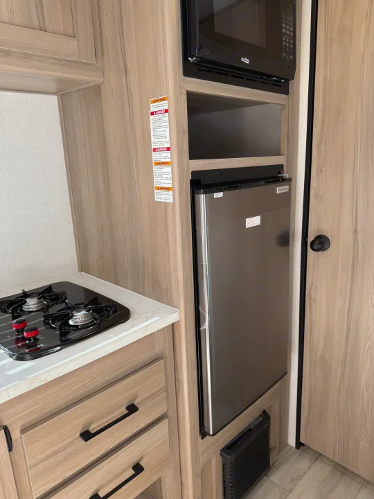 Compact RV rental kitchen featuring a two-burner stove, microwave, and refrigerator, set in light wood cabinetry.. Keystone Coleman 17B 2025