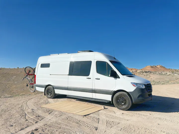 White camper van RV rental with bike on back, mat on ground, parked in sunny desert with hills.. Mercedes-Benz Sprinter 2021