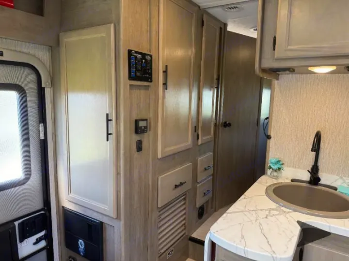 Inside a modern RV rental, observe the kitchen sink, light-toned cabinetry, and systems control panel near the entry door.. Coachmen RV Freelander 26DS 2023