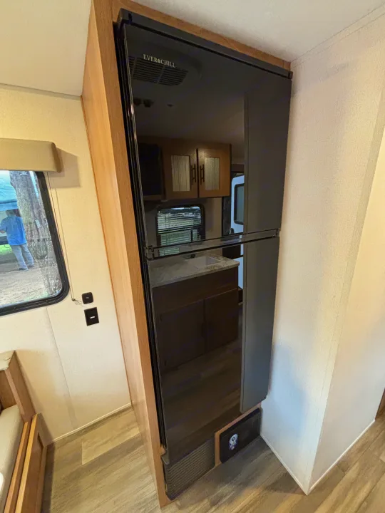 Modern RV rental interior: a large black refrigerator (Ever Chill) with reflective doors, wood trim, and a window.. Avenger 16RD 2024