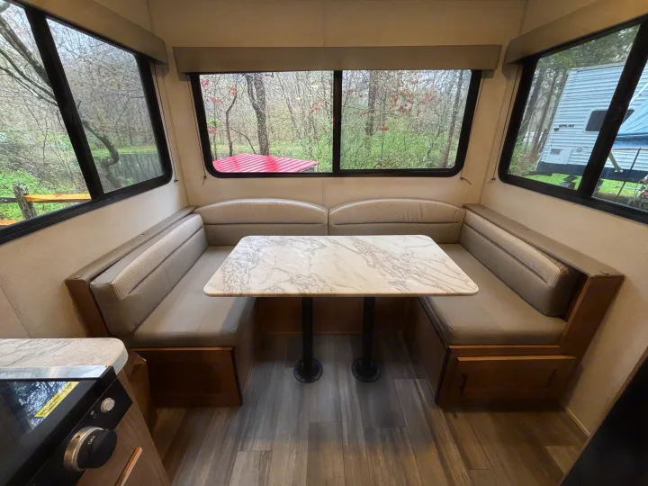 Inside an RV rental: a U-shaped dinette with faux marble table, large windows revealing nature & another RV.. Avenger 16RD 2024