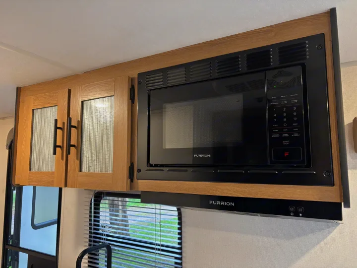 Furrion microwave and range hood in an RV rental kitchen, mounted above light wood cabinets with patterned glass doors.. Avenger 16RD 2024