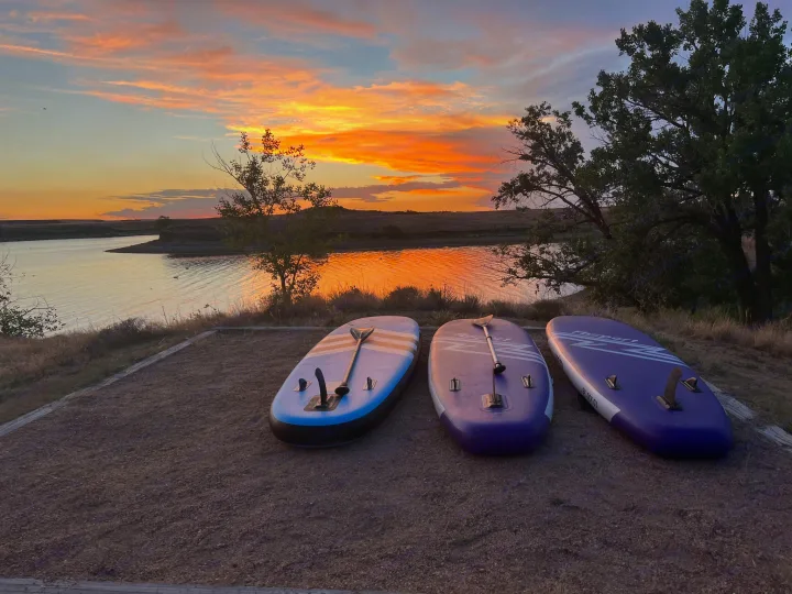 Three colorful paddleboards on the shore of a lake, reflecting a breathtaking sunset with vibrant orange and pink clouds. An ideal RV rental getaway.. Keystone RV Bullet Premier Ultra Lite 2022