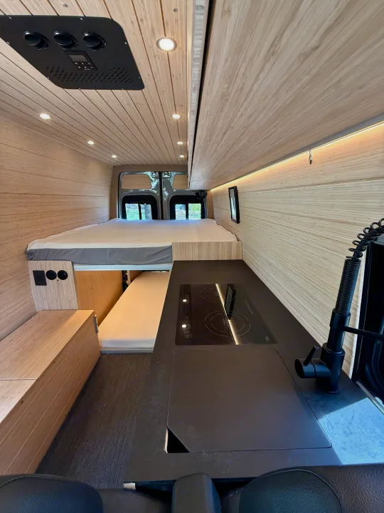 RV rental: Inside a modern camper van featuring a spacious bed, sleek kitchen with cooktop, and warm wood paneling.. Mercedes-Benz Sprinter 2024