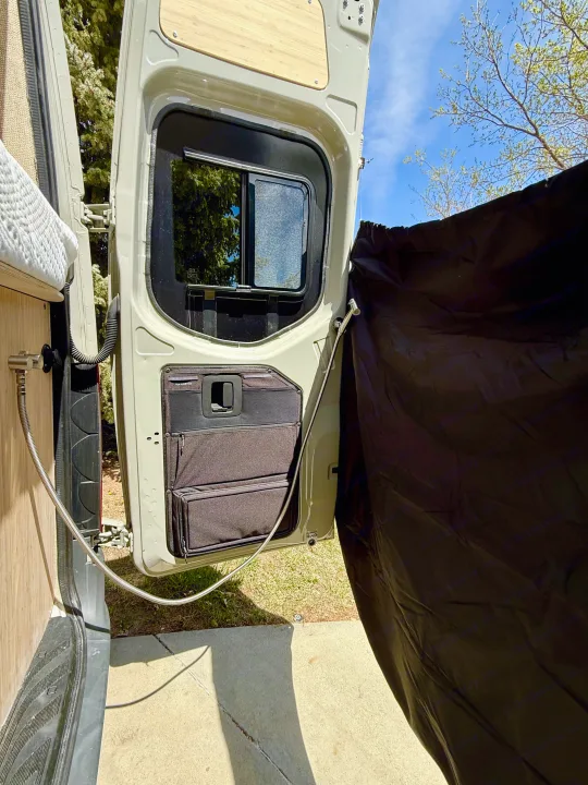 This RV rental detail shows a van's rear door open with built-in storage and an outdoor shower privacy curtain setup.. Mercedes-Benz Sprinter 2024