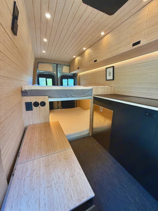 RV rental featuring a sleek camper van interior with natural wood paneling, an elevated bed, and compact kitchen.. Mercedes-Benz Sprinter 2024