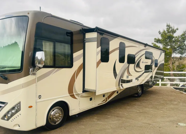 This image shows a cream & brown Class A motorhome, perfect for RV rental, parked with its slide-out extended, under a cloudy sky.. Thor Hurricane 2018