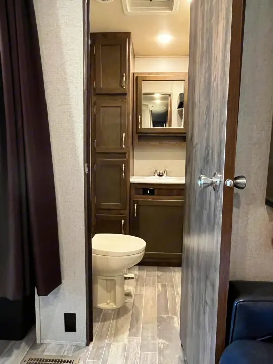Inside an RV rental: a compact bathroom featuring a white toilet, sink vanity, mirrored cabinet, and dark wood storage.. Dutchmen Astoria 2018