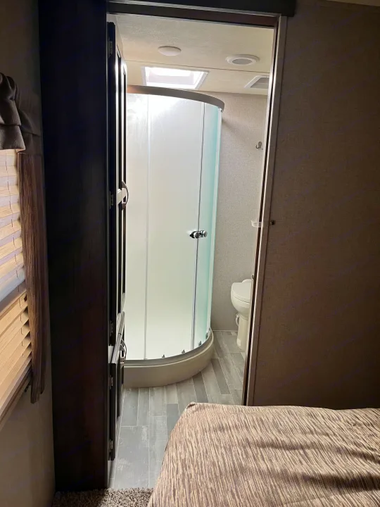 This RV rental bathroom features a curved glass shower and toilet, seen from the cozy bedroom. Note skylight & wood-look floor.. Dutchmen Astoria 2018