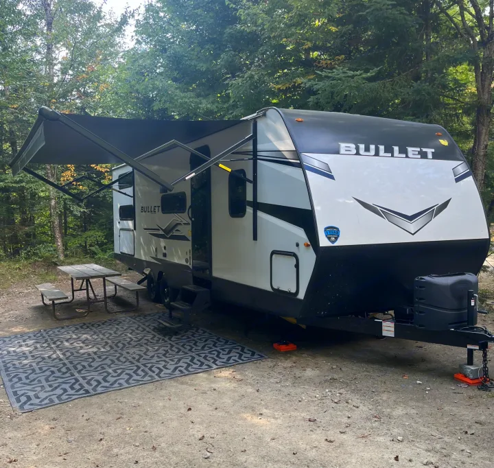 Cozy RV rental setup with extended awning and picnic area in a serene wooded campsite.. Keystone Bullet 2024