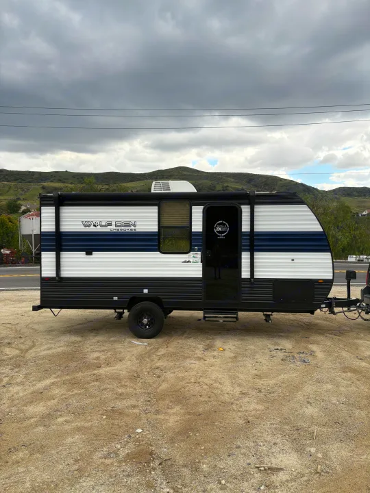 This sleek, blue, white, and black Wolf Den Cherokee RV rental is parked on a dirt lot, ready for your outdoor adventures.. Forest River Cherokee Wolf Den 16EV 2024
