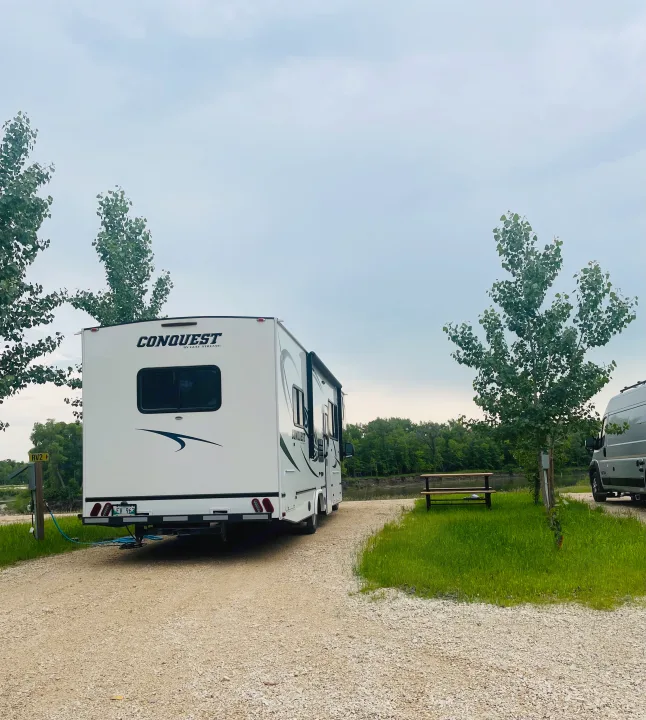 A white Conquest RV rental is set up at a gravel riverside campsite (RV2), ideal for a relaxing getaway.. Gulf Stream Conquest 2020