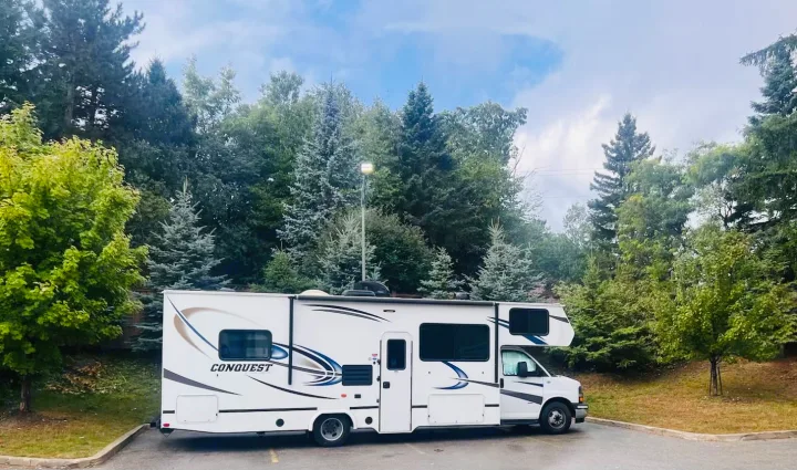 A white RV rental is parked on asphalt, surrounded by lush green and blue-green trees under a partly cloudy sky.. Gulf Stream Conquest 2020